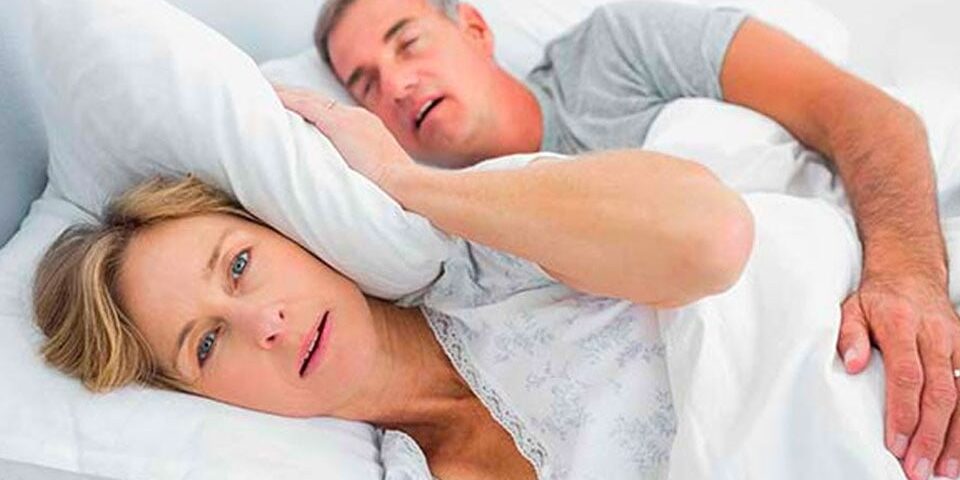 Man snoring loudly in bed while partner covers her ears with pillow and looks annoyed